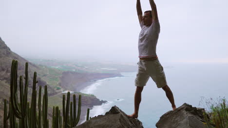 Engaging-in-sun-salutation-yoga-against-a-mountain-backdrop-with-ocean-views,-a-fit-young-man-embraces-meditation-and-exercise,-highlighting-a-sporty-and-healthy-lifestyle
